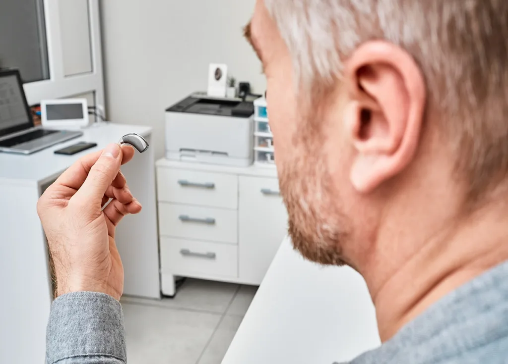 A middle aged man with graying hair sitting in a hearing clinic holding up a small over the ear hearing aid as if to inspect it for repair